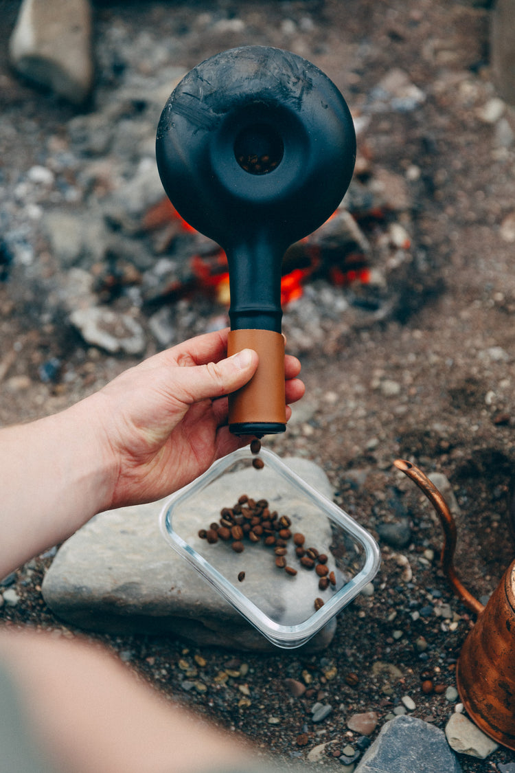Hand holding a black and brown device over a container of coffee beans near a fire.